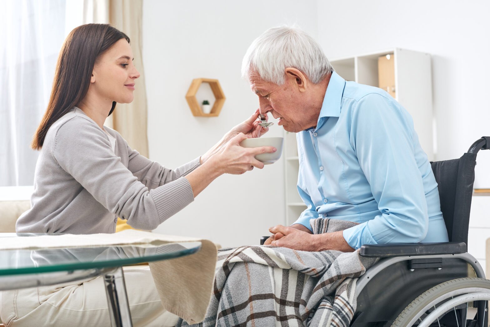 Young Social Worker Holding Bowl With Soup And Spoon By Mouth Of Senior Disable Man Sitting In Wheelchair While Helping Him With Eating